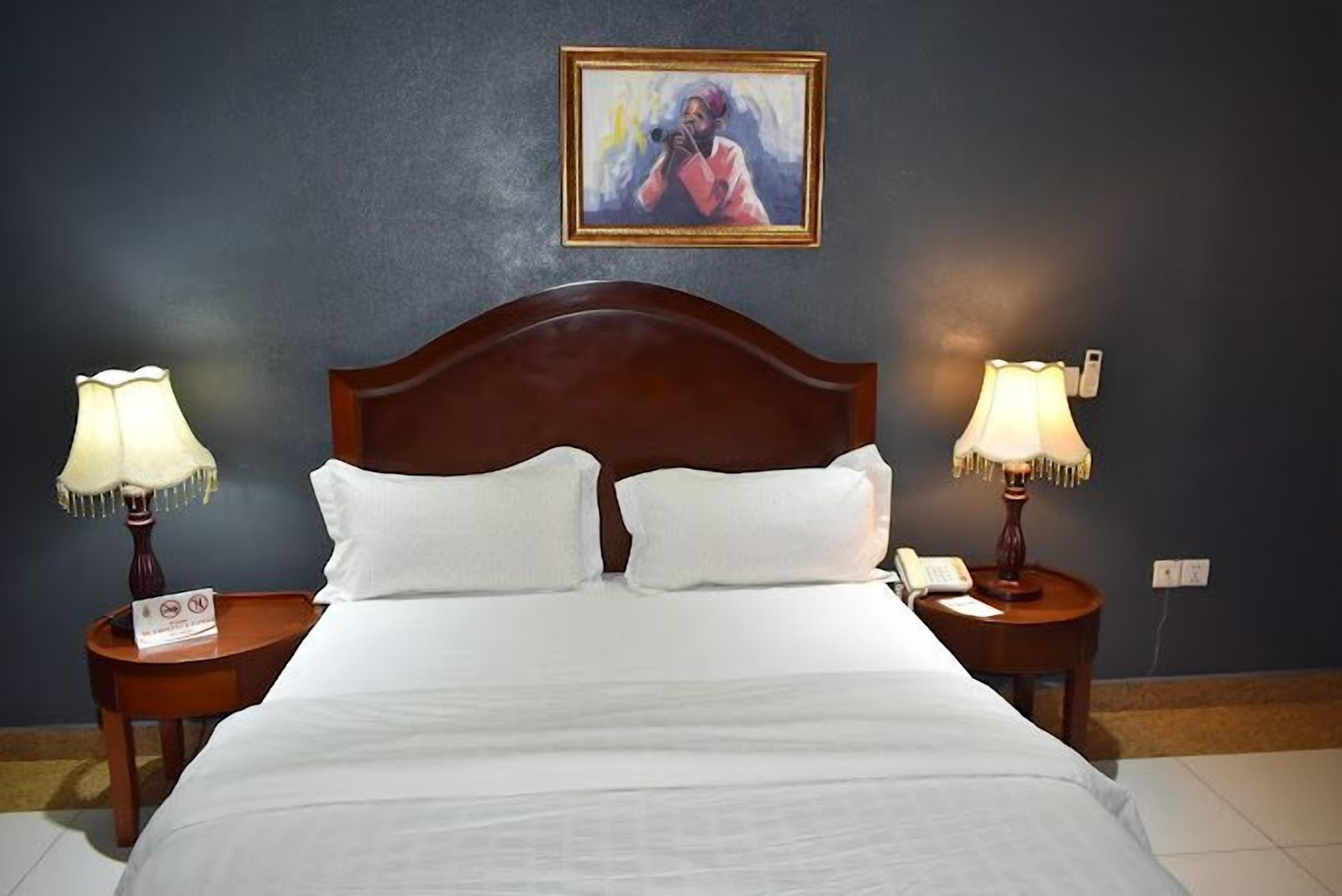 A neatly made bed with white linens, two bedside tables with lamps, a painting on the wall, and a telephone on one of the tables.