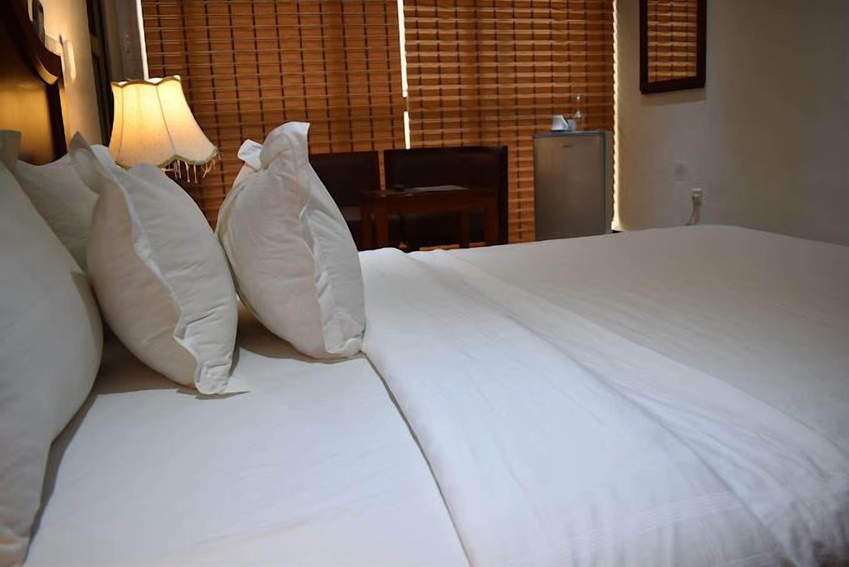 A neatly made bed with white linens and pillows, a bedside lamp, and a wooden headboard.