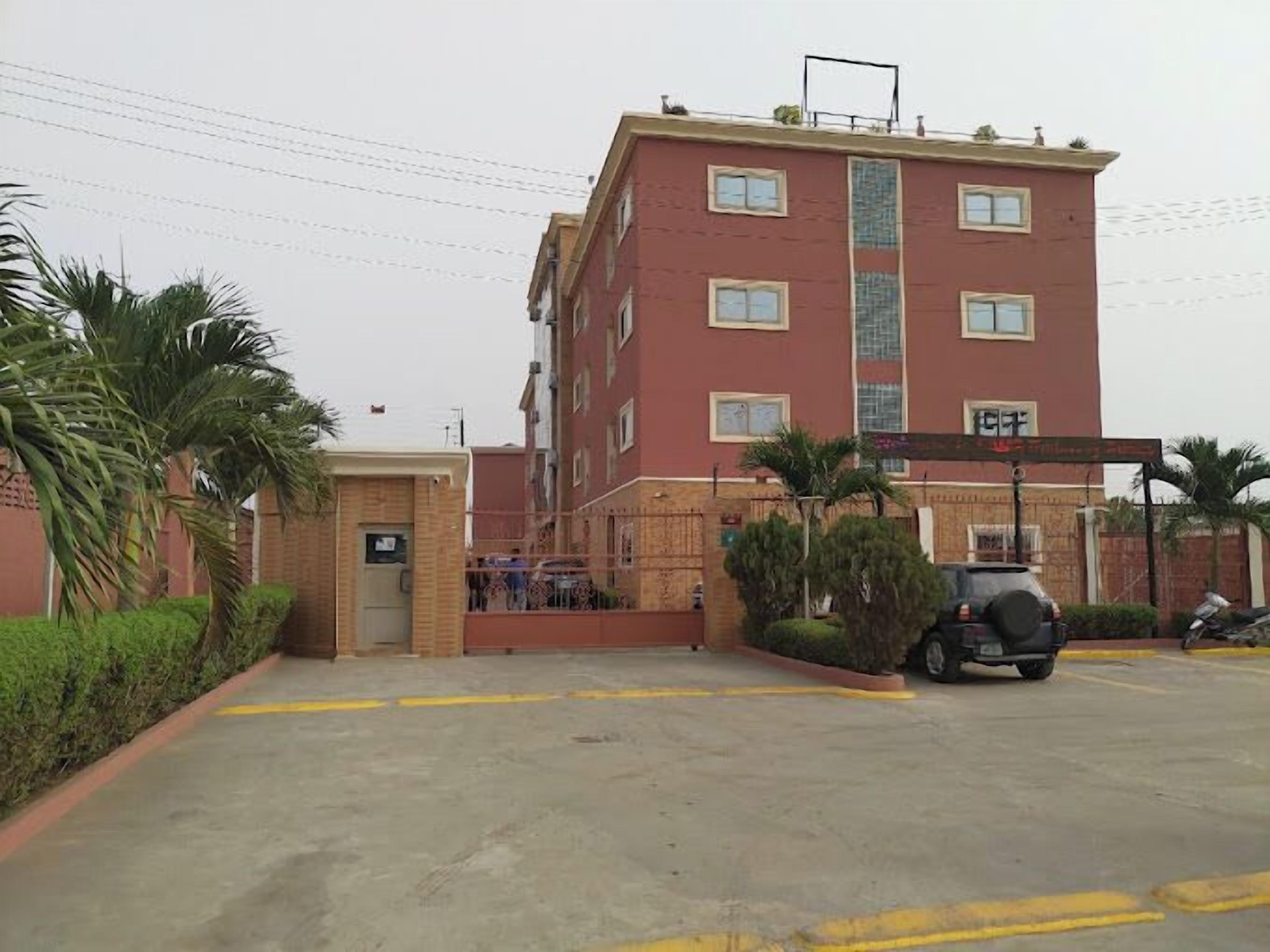 A multi-story building with a red facade, a gated entrance, and a parking area with a few vehicles.