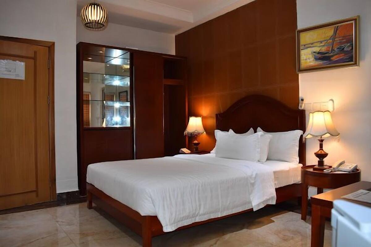 A hotel room with a large bed, wooden headboard, two bedside lamps, a nightstand with a phone, a mirror, and a painting on the wall.