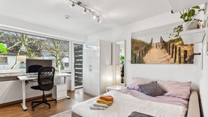 Une chambre moderne avec un lit, un bureau et un ordinateur. Il y a une fenêtre donnant sur des arbres et un immeuble.