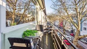 Un balcon avec une petite table et des chaises, donnant sur une rue bordée de voitures garées et d’immeubles.