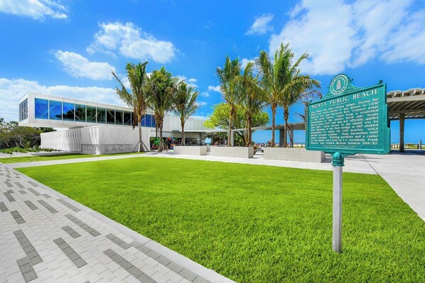 A modern building with a glass facade, a grassy area, palm trees, and a sign about Siesta Beach history.