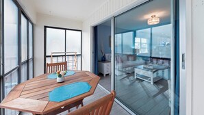 Uma varanda com mesa e cadeiras de madeira, e porta de vidro que dá para uma área de estar interna com sofá e mesa de centro.