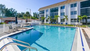 Uma piscina com um edifício branco ao fundo, área cercada com espreguiçadeiras e céu claro.