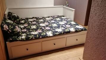 Ein Bett mit blumigem Überwurf, einem Holzkopfende und einer weißen Wand mit Regal darüber.