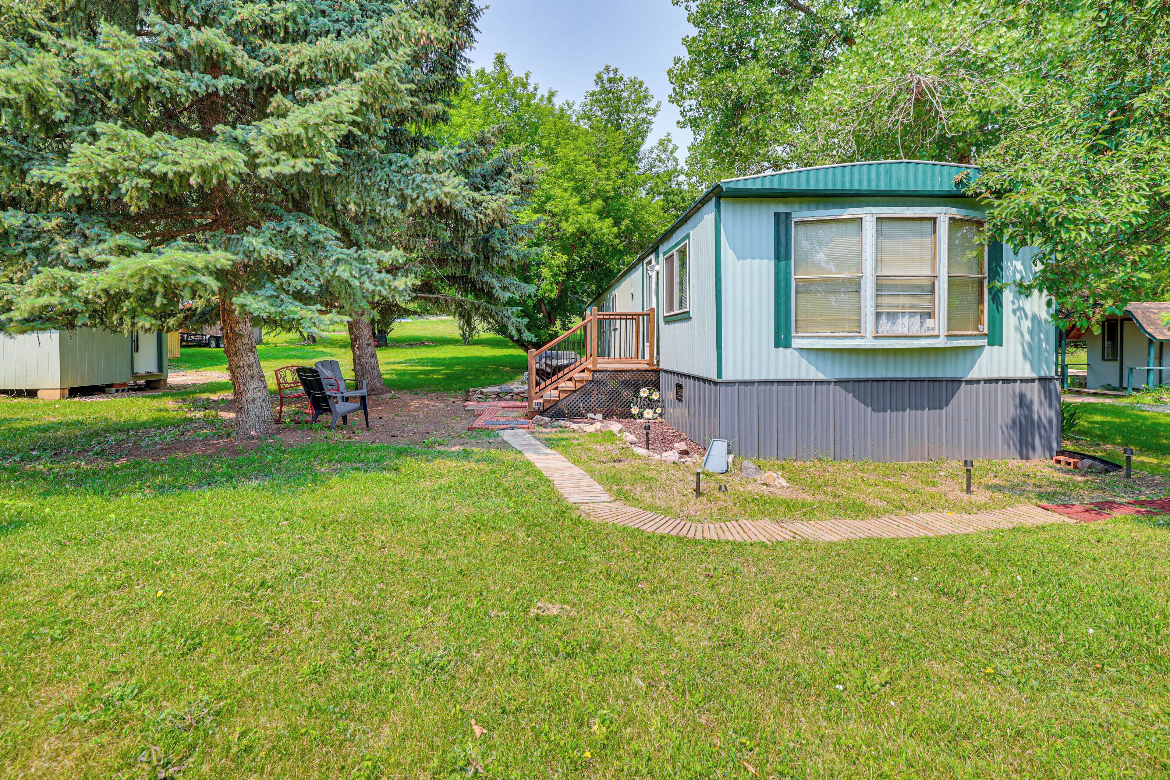 A small, single-story mobile home with a porch and a small deck, surrounded by trees and grass.