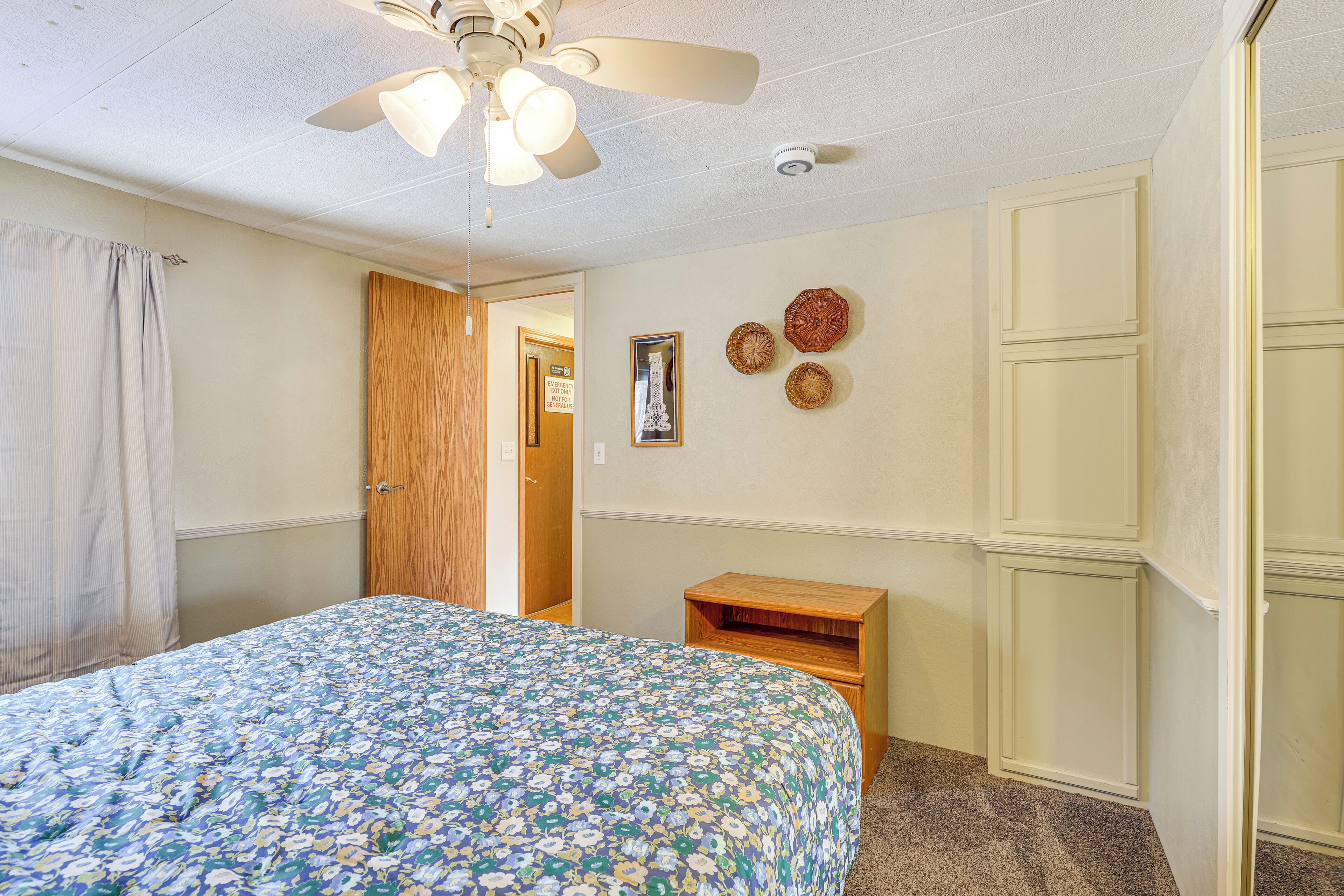 A bedroom with a bed, a ceiling fan, a wooden door, a small wooden nightstand, and a wall with decorative plates.
