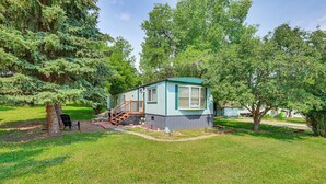 A single-story mobile home with a small porch and a covered outdoor area, surrounded by trees and grass. - 2 Mi to Sturgis Motorcycle Rally: Peaceful Home (Sturgis)