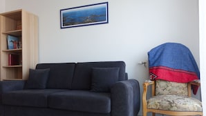 Un salón con un sofá de color gris oscuro, una estantería de madera, un cuadro en la pared y un sillón con un cojín estampado.