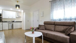 Una sala de estar moderna con un sofá marrón, una mesa de centro blanca con un jarrón de flores, y una cocina al fondo.