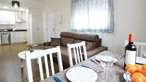 Una cocina moderna con armarios blancos, una zona de comedor con mesa para dos personas y un sofá.