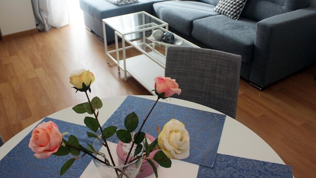Un salon moderne avec un canapé gris, une table basse en verre et un vase rempli de roses, posé sur une nappe à motifs.