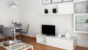 Un salon moderne avec un meuble de rangement blanc pour l’équipement audiovisuel, un téléviseur à écran plat, une table basse en verre et un coin repas avec une petite table et des chaises.