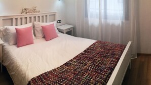 Une chambre avec un lit blanc, des oreillers roses, une table de chevet blanche, une lampe fixée au mur et une fenêtre ornée de rideaux légers.
