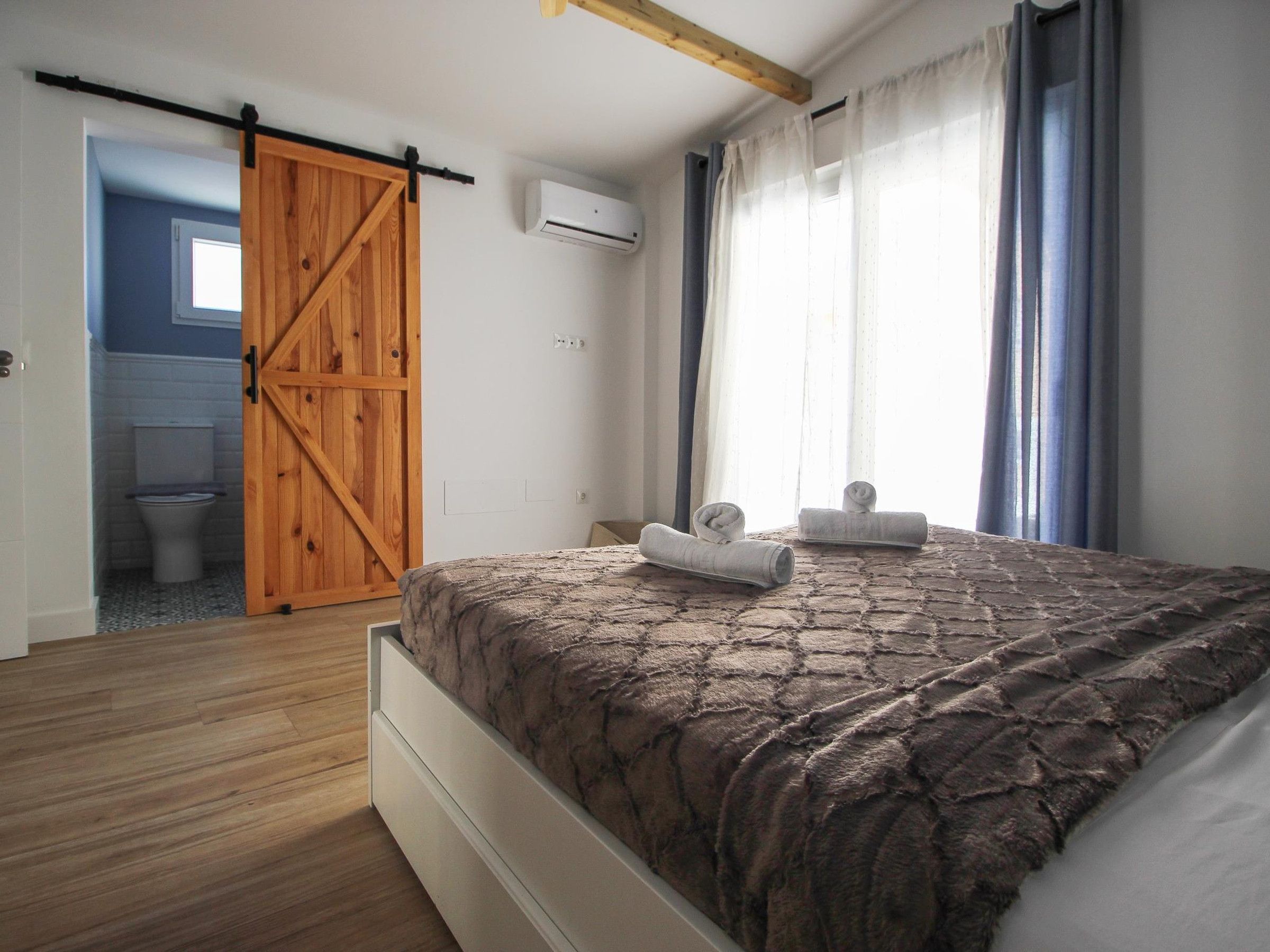 A bedroom with a wooden door, a bed with a patterned bedspread, a window with curtains, and a view of a bathroom.