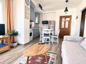 A compact living space with a sofa, a dining table, a washing machine, a TV, and a small kitchen area.