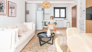 Una sala moderna con un sofá blanco, una mesa de centro redonda y una silla de mimbre. Se ve una cocina al fondo con una ventana y una lámpara colgante.