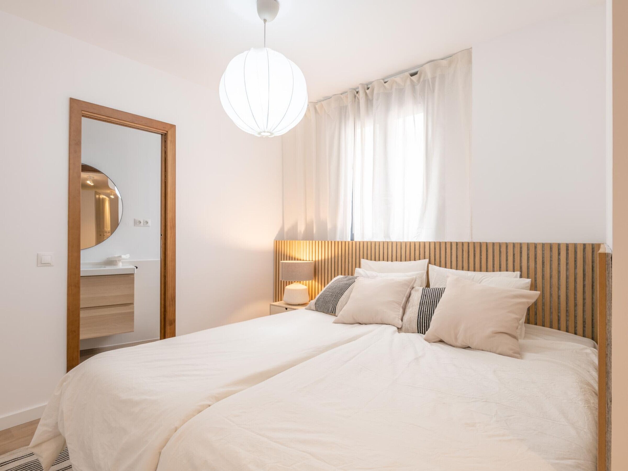 A hotel room with a large bed, a bedside table with a lamp, a mirror, and a window with curtains.