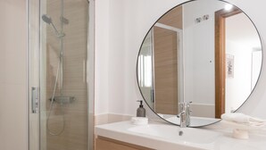 Un baño moderno con ducha envuelta en vidrio, espejo redondo y lavamanos blanco sobre tocador de madera.