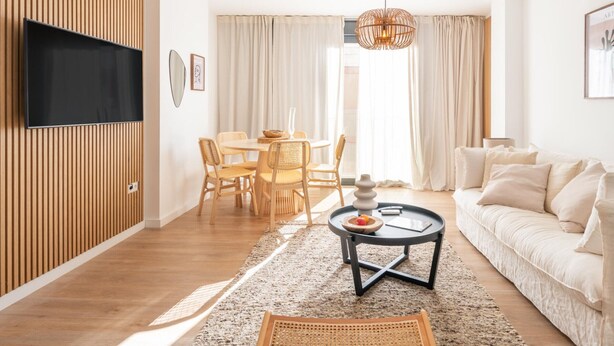 Una sala moderna con un televisor de pantalla plana montado en una pared de paneles de madera, una mesa de comedor redonda con sillas de mimbre, una mesa de centro negra con un tazón y una vela, y un sofá color beige.