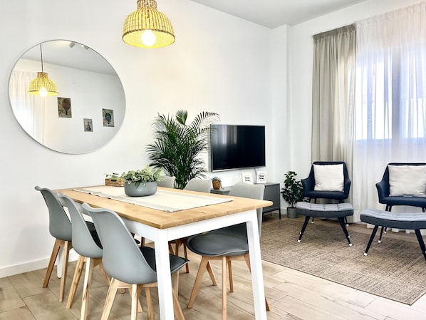 A modern living room with a wooden dining table, grey chairs, a large mirror, a television, and a potted plant.