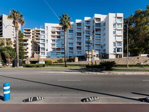 Ein mehrgeschossiges Wohngebäude mit Balkonen, eine gepflasterte Straße im Vordergrund und Palmen.