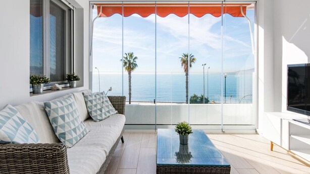 Una sala de estar moderna con un sofá, una mesa de centro de vidrio y un televisor. Hay un ventanal grande con un toldo naranja y vista al mar.