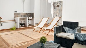A modern, well-lit room with a tiled floor, a small kitchenette, two lounge chairs, a sofa, and a potted plant.