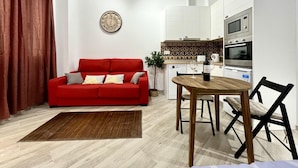 A compact living space with a red sofa, a dining table with two chairs, a kitchen area with modern appliances, and a decorative wall clock.
