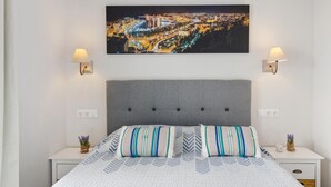 Un dormitorio ordenado con vistas a la ciudad, un cabecero tapizado en gris y dos lámparas de noche.