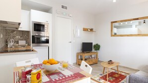Una cocina moderna con horno empotrado y microondas, un comedor con mesa y sillas de madera, y un televisor pequeño sobre un mueble de madera.