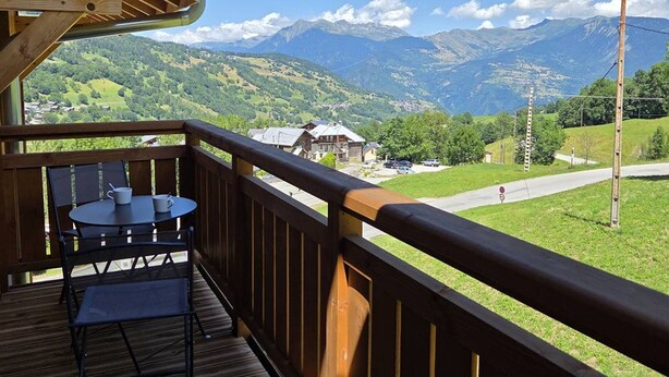 Una terraza de madera con una mesa y sillas, con vista a un paisaje montañoso que incluye casas y una carretera.