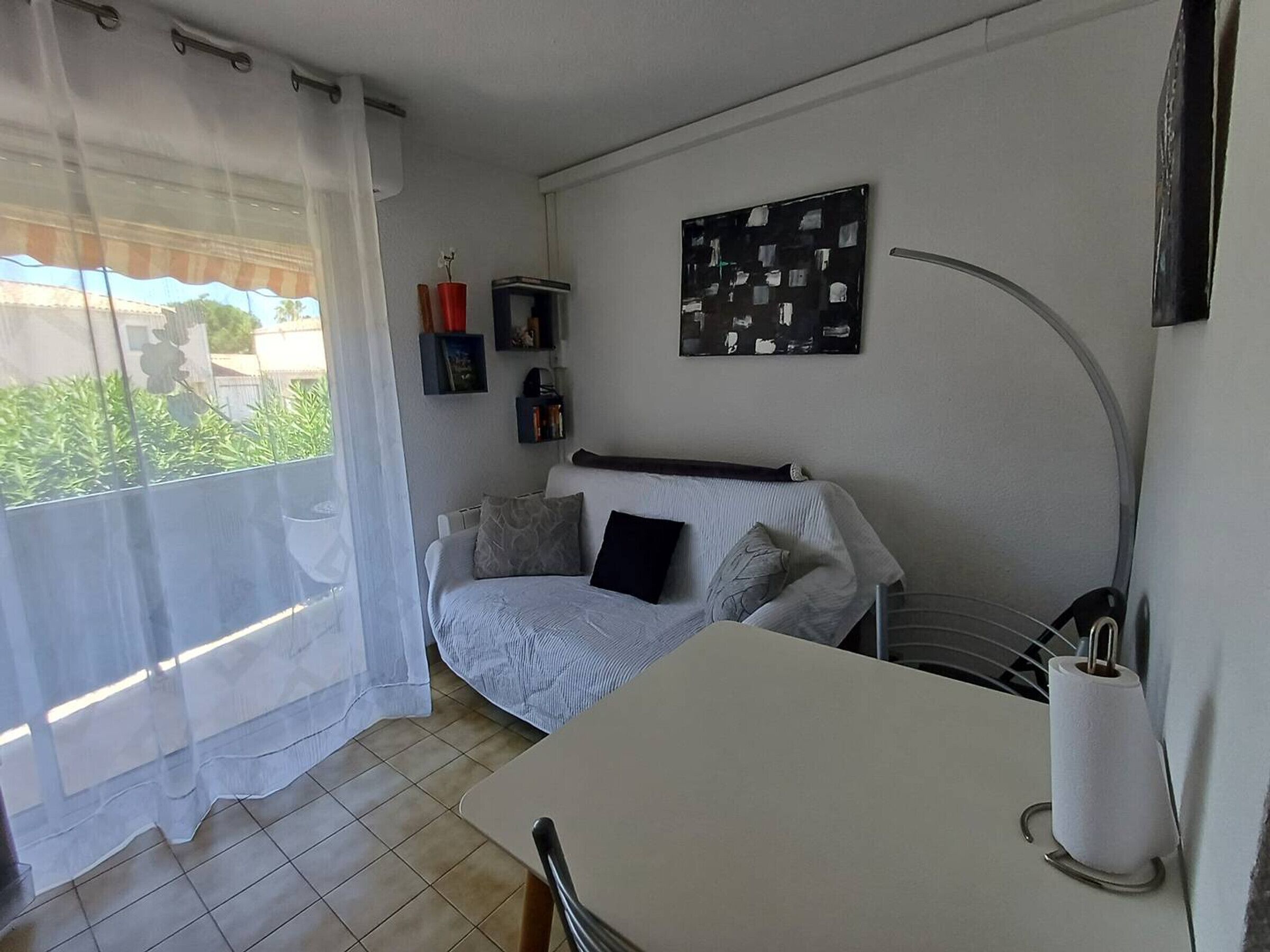 Una habitación pequeña y bien iluminada, con una cama, una mesa y una silla. Se aprecia una ventana con cortinas de encaje, un cuadro en la pared y un estante con objetos.