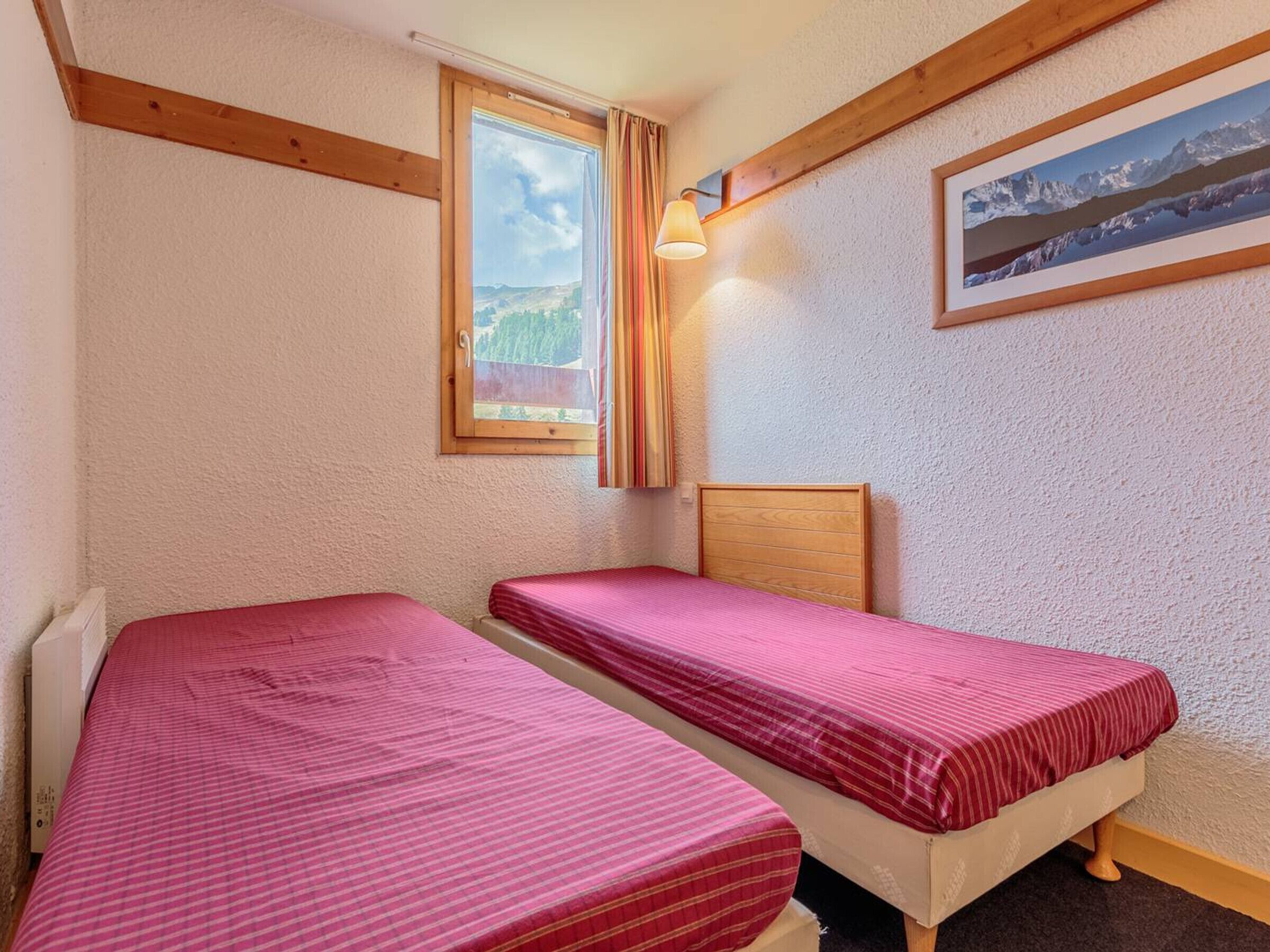 A small room with two single beds, a window with curtains, a mounted picture, and a wooden beam ceiling.