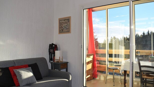 A living room with a grey sofa, a small table with a candle, and a sliding glass door leading to an outdoor area with a wooden deck and a table. - Cozy 2-Room Apartment in Bolquère, Near Shops and Skiing (Bolquère-Superbolquère)