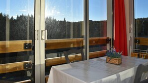 A dining area with a table set for two, large windows with a view of a forest, and a red curtain on the right. - Cozy 2-Room Apartment in Bolquère, Near Shops and Skiing (Bolquère-Superbolquère)