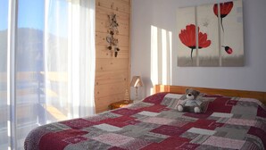 A bedroom with a large bed, a wooden headboard, a window with sheer curtains, a wall-mounted decoration, and a painting of red flowers. - Cozy 2-Room Apartment in Bolquère, Near Shops and Skiing (Bolquère-Superbolquère)