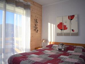 A bedroom with a large bed, a wooden headboard, a window with sheer curtains, a wall-mounted decoration, and a painting of red flowers. - Cozy 2-Room Apartment in Bolquère, Near Shops and Skiing (Bolquère-Superbolquère)