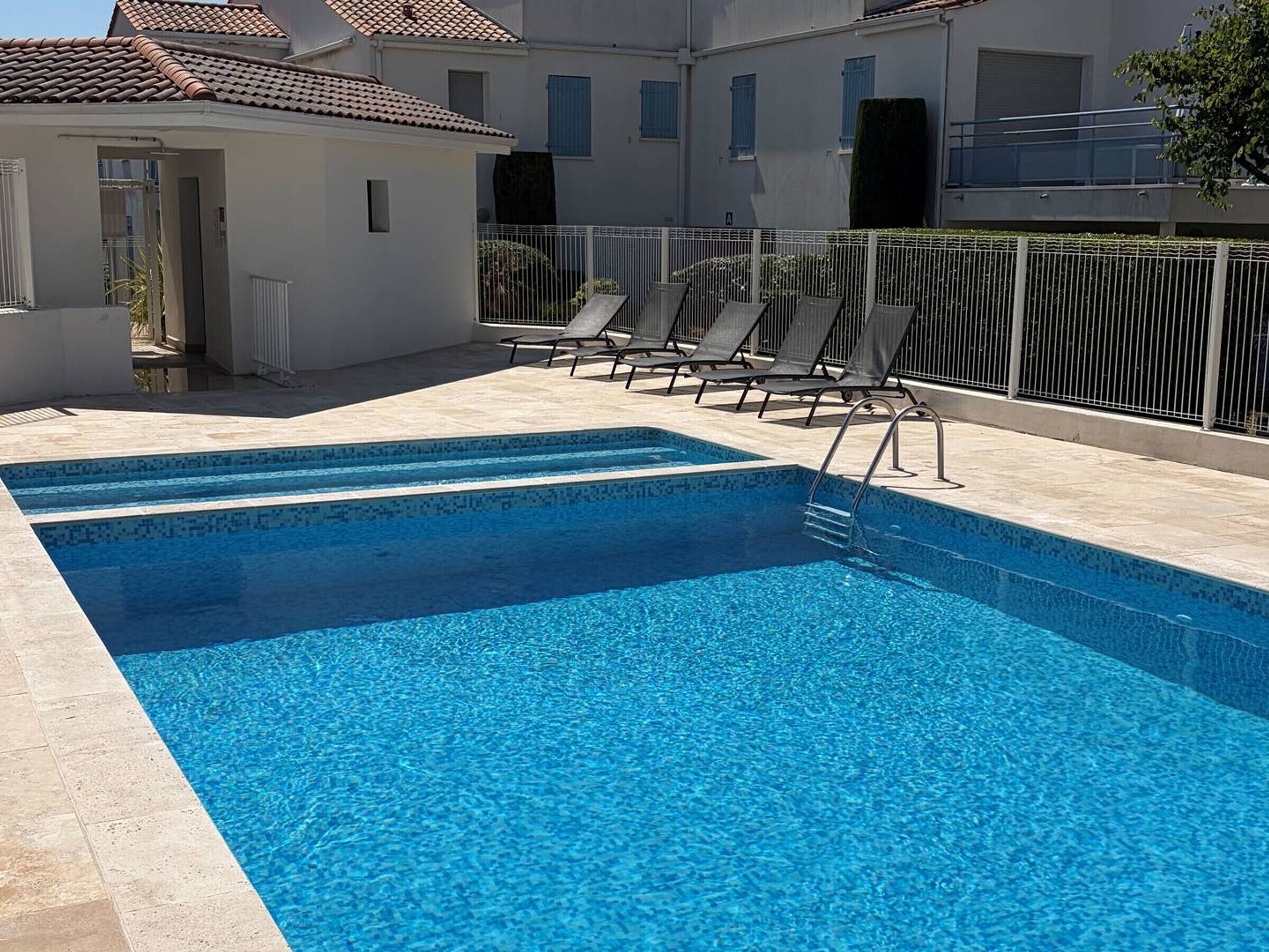 Une piscine rectangulaire à l’eau d’un bleu cristallin, entourée d’une terrasse carrelée de couleur blanche. Plusieurs chaises longues sont alignées le long du bord de la piscine.