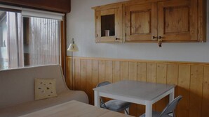 A room with a wooden wall panel, a white table with two chairs, a sofa with a patterned pillow, and a window with blinds. - Cozy 2-room apartment with garden in Bolquère (Bolquère-Pyrénées 2000)