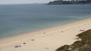 Une plage avec un rivage sablonneux, quelques personnes qui prennent un bain de soleil et une colline verdoyante à l’horizon.