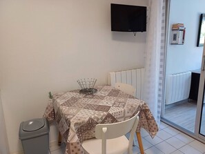 Un petit coin repas avec une table recouverte d’une nappe à motifs, une chaise, un téléviseur fixé au mur et un radiateur.