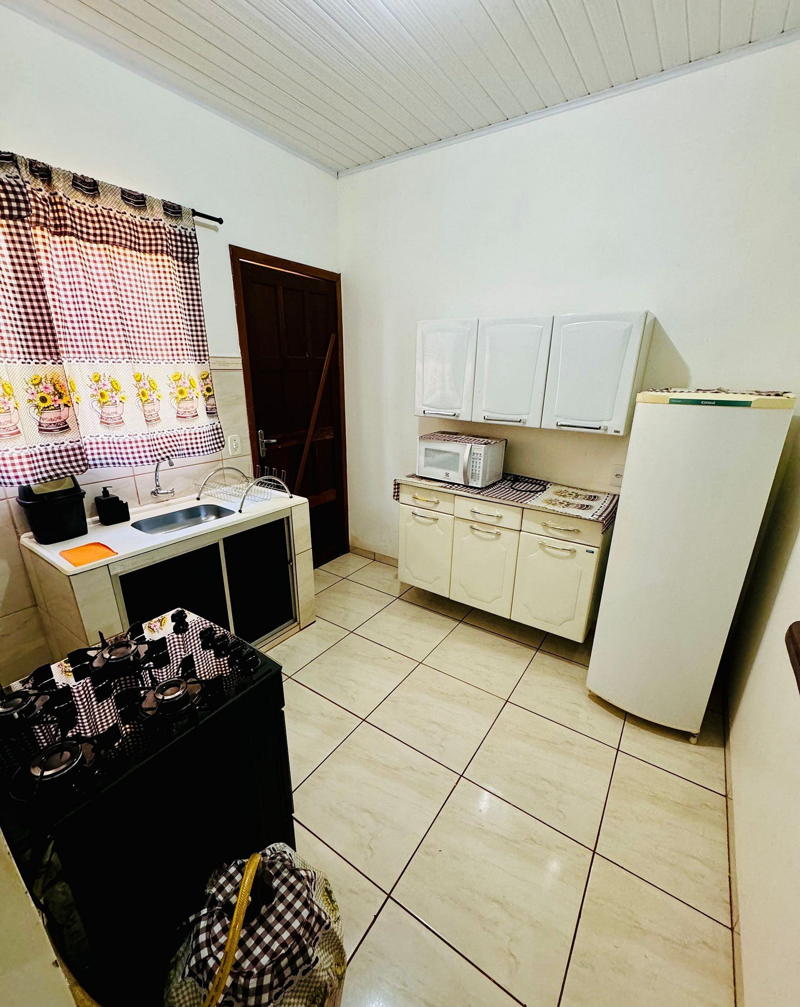 A small kitchen with a gas stove, a microwave, a refrigerator, and a sink. There is a window with a checkered curtain, and a door leading to another room.