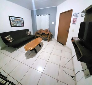 A living room with a sofa, a wooden bench, a television, and a door with a sign. - Casa Flor do Ipê Homes  (Nobres)