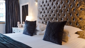 Une chambre d’hôtel avec une tête de lit capitonnée, une table de chevet blanche avec une lampe et deux oreillers foncés.