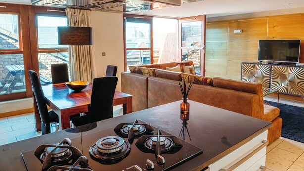 Une cuisine moderne dotée d’un îlot central, un coin repas avec une table et des chaises en bois, et un espace de vie avec un canapé et un téléviseur.