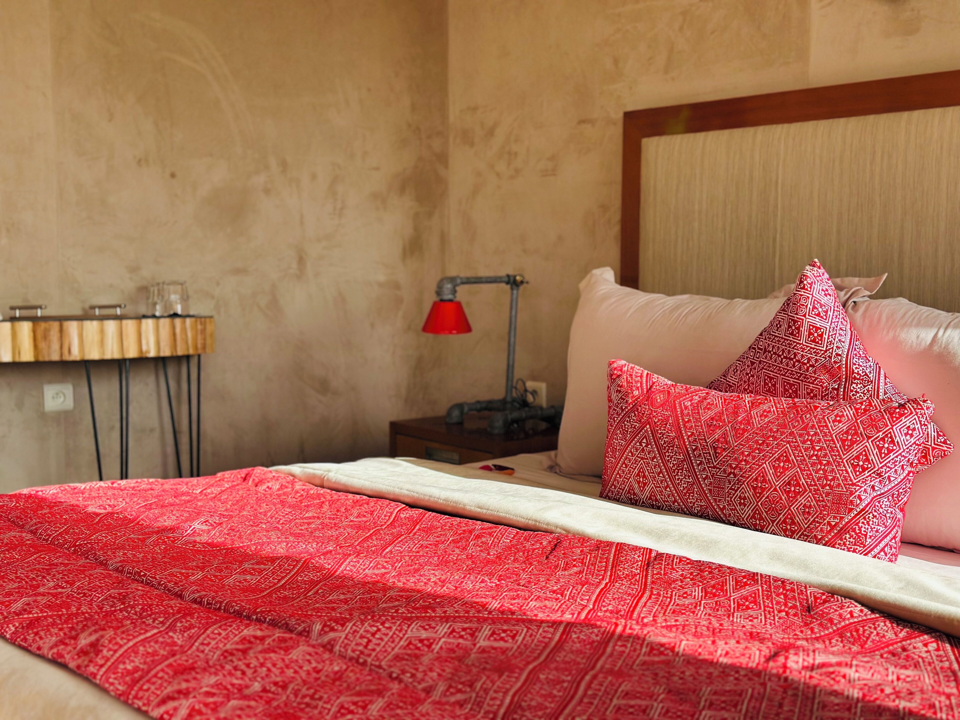 A neatly made bed with a red patterned blanket, a red lamp on a bedside table, and a wooden side table with a glass container.