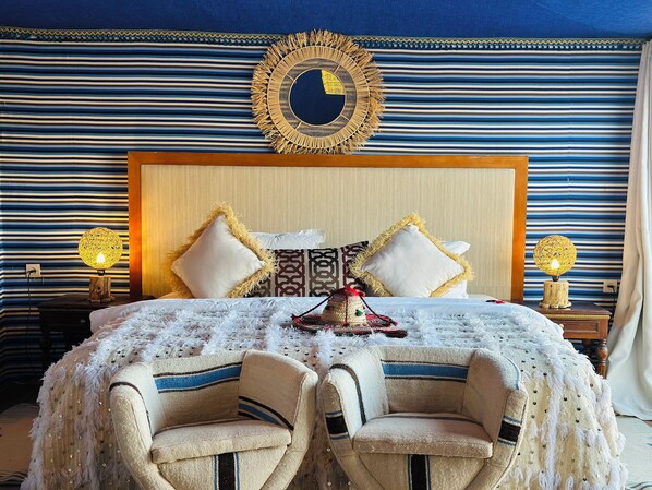 Habitación de hotel con pared azul y blanca a rayas, cabecera de madera, dos almohadas blancas, un espejo decorativo y una mesa con mantel blanco y dos sillas.