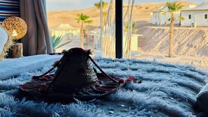 Ein Bett mit einem gewobenen Kopfteil und einer Lampe auf einem flauschigen Teppich, mit Blick auf eine Wüstenlandschaft mit Zelten und Palmen.